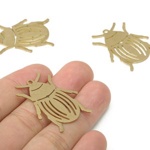 Pode incluir: Três pingentes de metal dourado em forma de besouro. Os pingentes são detalhados com desenhos recortados e pequenos orifícios para fixar em joias ou outros trabalhos artesanais. Os pingentes de besouro são mostrados em um fundo branco.