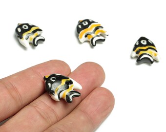 May include: Small, decorative fish-shaped beads. Each bead features a black, yellow, and white design. The beads are shown on a white background, with one bead held in a person's hand. These beads could be used for jewelry making or other craft projects.