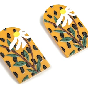 May include: A pair of yellow, arched earrings with a black spotted design. Each earring has a white daisy with a yellow centre, green leaves, and brown stems. These earrings are a distinctive accessory.