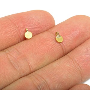 Può includere: Due piccoli pendenti rotondi color oro con un anello nella parte superiore, appoggiati sulle dita di una persona. I pendenti hanno un design semplice e minimalista, adatti per la creazione di gioielli o l'artigianato.