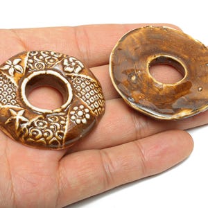 May include: Two brown ceramic donut-shaped beads. One bead has a detailed white floral and geometric pattern. The other bead is a solid brown colour. Both beads have a hole in the centre.