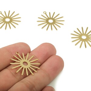 Puede incluir: Cuatro dijes dorados en forma de sol con un agujero central y líneas radiales. Un dije se sostiene en una mano, mostrando el diseño. Adecuado para la fabricación de joyas y proyectos de artesanía.