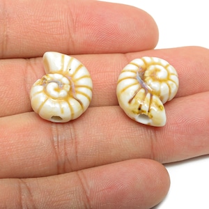 Pode incluir: Duas contas de cerâmica em forma de concha, cada uma com um desenho em espiral. As contas são brancas com detalhes em castanho e bege. As contas são seguradas numa mão.
