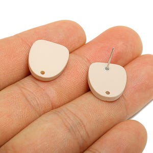 Puede incluir: Dos pendientes de botón beige con forma redondeada y asimétrica. Cada pendiente tiene un pequeño agujero y un poste plateado para perforar. Los pendientes se muestran en una mano sobre un fondo blanco.