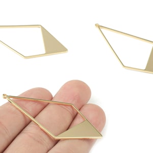 Könnte beinhalten: Goldfarbene geometrische Anhänger. Jeder Anhänger hat eine rautenförmige Form mit einem dreieckigen Abschnitt. Die Anhänger haben eine kleine Schlaufe zum Befestigen an Schmuck. Das Bild zeigt drei Anhänger, einer wird von einer Hand gehalten.