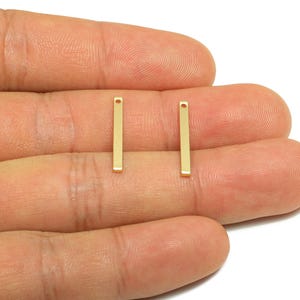 May include: Two gold-colored rectangular bar charms with a small hole at the top. These jewelry findings are shown against a neutral background. The charms are small, measuring approximately 0.75 inches in length.