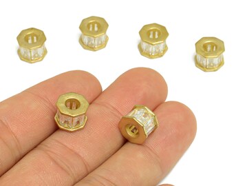 Puede incluir: Seis cuentas doradas, octagonales, con inserciones de cristal transparente facetado. Las cuentas se muestran sobre un fondo blanco, algunas sujetas en una mano. Estas cuentas son probablemente para la fabricación de joyas o manualidades.