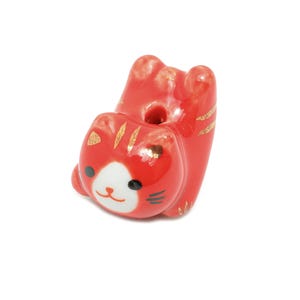 Może przedstawiać: Czerwona ceramiczna figurka kota ze złotymi akcentami. Kot ma białą twarz z czarnymi wąsami i oczami. Figurka jest w zabawnej pozie. Przedmiot dekoracyjny.