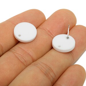 May include: Two white, circular stud earrings with a post and a small hole. The earrings are held in a hand, displaying the front and back. The earrings have a simple, minimalist design.