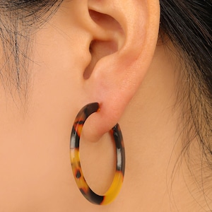 May include: A close-up of a tortoiseshell hoop earring worn on an ear. The earring is a circular shape with a gradient of colors, including black, brown, and yellow. The earring is made of a glossy material.
