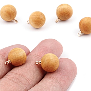 May include: A collection of wooden round beads with a light brown color. Each bead has a small gold-colored loop attached. Two beads are held in a person's hand. The beads are likely for jewelry making or craft projects.