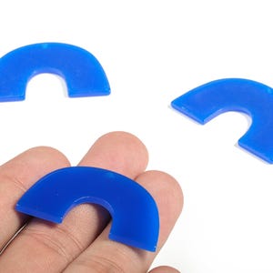Pode incluir: Três pingentes de acrílico azul brilhante em forma de semicírculo. Um pingente é segurado em uma mão, enquanto dois outros são posicionados acima. Os pingentes têm um acabamento liso e brilhante e um pequeno orifício na parte superior para enfiar.