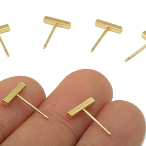 May include: Gold-colored bar stud earrings. The earrings have a simple, minimalist design with a rectangular bar and a thin post. Several pairs are displayed, with two held between fingers.