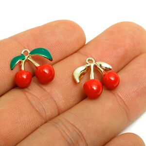 Op de afbeelding: Twee kersvormige bedels. Elke bedel heeft twee rode kersen met groene bladeren en een goudkleurige steel. De bedels zijn klein en waarschijnlijk bedoeld voor sieraden of knutselprojecten.