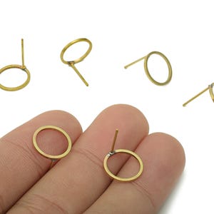 May include: Several small, gold-colored earring posts with circular hoops are displayed. The earrings are scattered across a white surface, with some held by a hand. The earrings are made of a thin metal and have a simple, minimalist design.