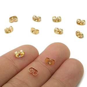 May include: Gold-colored earring backs in various shapes and sizes are scattered on a white surface. Several are butterfly-shaped, while others are a simple post-style. Some are held between fingers, showcasing the different designs.