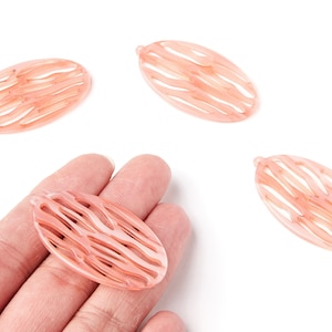 Peut inclure: Quatre perles ovales en résine pêche avec un motif ajouré texturé. Une perle est tenue dans une main, tandis que les autres sont dispersées sur une surface blanche. Chaque perle a un petit trou en haut pour l'enfilage.