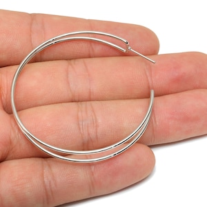 May include: Silver-toned hoop earrings with a double-band design. The earrings are held in a hand against a white background. The hoops are circular and have a simple, elegant style, suitable for everyday wear.