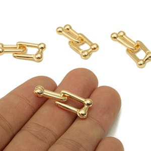 May include: Gold-colored metal jewelry findings in a U-shaped design. The findings have a polished finish and are shown in a close-up view. The image shows several pieces, with one held in a hand.