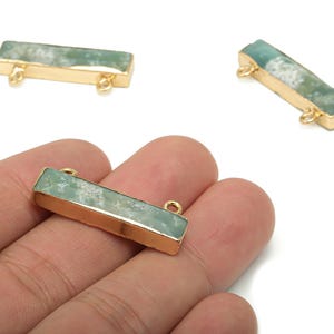 Puede incluir: Tres colgantes rectangulares de piedras preciosas con marcos de metal dorado. Las piedras preciosas tienen un aspecto veteado en verde claro y blanco. Cada colgante tiene dos pequeños bucles para sujetar. Un colgante se sostiene en una mano.