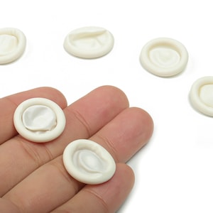 Puede incluir: Una colección de pequeños dediles de color blanquecino, algunos sobre una mano. Los dediles son circulares con un centro ligeramente elevado, y están hechos de un material suave y flexible. Están diseñados para proteger los dedos.
