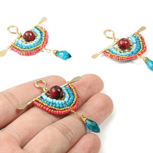 May include: Handmade beaded earrings in a unique design. Each earring features a fan-shaped element with turquoise, red, and gold beads, a red bead center, and a dangling blue teardrop. The earrings have a gold-colored metal finish.
