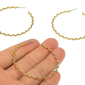 Peut inclure: Deux boucles d'oreilles créoles dorées avec un motif ondulé. Les boucles d'oreilles sont présentées sur fond blanc. Les boucles d'oreilles sont tenues par une main.