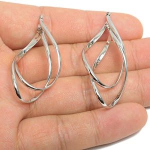 May include: A pair of silver-colored, teardrop-shaped earrings. Each earring features a double-loop design, with the loops intertwined. The earrings are displayed against a light background, highlighting their reflective surface and intricate form.