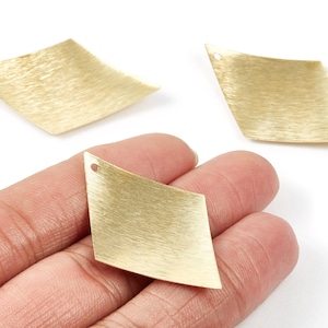 May include: Three gold-coloured, diamond-shaped metal jewellery components. One is held in a hand, and two are on a white surface. Each component has a small hole at the top for attaching to other jewellery pieces. The metal has a brushed texture.
