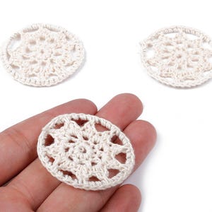 May include: Three off-white, crocheted doilies. One is held in a hand, while two others are placed on a white surface. The doilies have an open, circular design with a detailed, floral-like pattern. The crochet work appears delicate and handmade.
