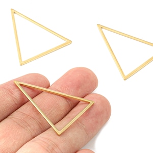 Peut inclure: Trois apprêts de bijoux dorés en forme de triangle. L'un est tenu dans une main, tandis que les deux autres sont dispersés sur un fond blanc. Ces composants géométriques sont idéaux pour la création de boucles d'oreilles, de pendentifs ou d'autres bijoux.