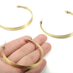 Puede incluir: Pendientes de aro dorados. Cada pendiente presenta un diseño de múltiples hebras, creando un efecto en capas. Los pendientes se presentan sobre un fondo blanco, resaltando su brillo metálico. Los aros se sostienen en una mano.