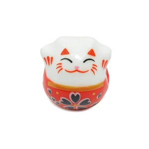 Może przedstawiać: Biała ceramiczna figurka Maneki-Neko, czyli kota szczęścia, z czerwonymi i złotymi akcentami. Kot siedzi z uniesioną prawą łapą, tradycyjny gest na szczęście i fortunę. Figurka jest ozdobiona czarnymi i różowymi wzorami kwiatowymi.