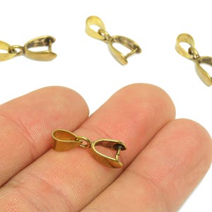 Könnte beinhalten: Nahaufnahme von mehreren kleinen, goldfarbenen Schmuckverschlüssen. Ein Verschluss wird zwischen den Fingern gehalten, mit drei weiteren im Hintergrund verstreut. Die Verschlüsse haben eine Schlaufe und ein gebogenes, offenes Design, geeignet für die Schmuckherstellung.