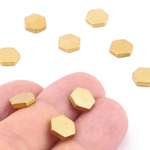 Puede incluir: Cuentas hexagonales de metal dorado sobre un fondo blanco. Algunas cuentas se sostienen en una mano. Estas pequeñas cuentas geométricas planas probablemente se utilizan para la fabricación de joyas u otros proyectos de artesanía.