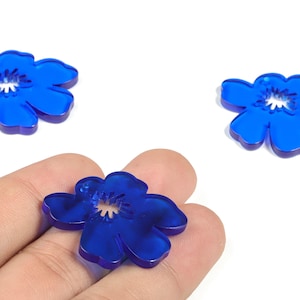 Op de afbeelding: Drie doorschijnende blauwe bloemvormige knutselversieringen. Eén wordt in een hand gehouden, terwijl de andere twee op een witte achtergrond verspreid liggen. De bloemen hebben vijf bloemblaadjes en een wit midden.