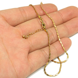 May include: A delicate gold-colored necklace with a rectangular link chain. The necklace is displayed on a hand, showcasing the adjustable length and clasp. The jewelry is likely made of a gold-toned metal.