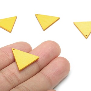 May include: A hand holding a yellow wooden triangle pendant with a small hole at the top. Three more yellow triangles are on a white background. These pendants are suitable for jewellery making.