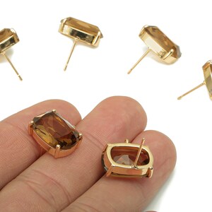 May include: Gold-toned stud earrings with rectangular, amber-colored gemstones. The earrings are displayed on a white background, with some held in a hand. The gemstones have a faceted cut, catching the light.