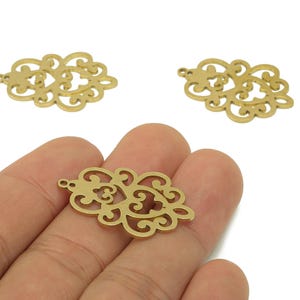 May include: Three gold-colored filigree charms with an ornate, openwork design. The charms have a small hole at the top for stringing. One charm is held in a hand, while the other two are on a white background.