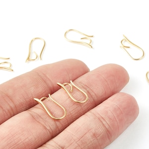 May include: Several gold-colored earring hooks are displayed against a white background. Two hooks are held by a hand. The hooks have a curved design, suitable for jewelry making or earring replacement.