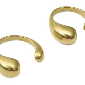 May include: Two gold-colored, open-ended rings. Each ring has a smooth, teardrop-shaped design on one end, curving around to an open, rounded tip. The rings are set against a white background, highlighting their polished, metallic finish.