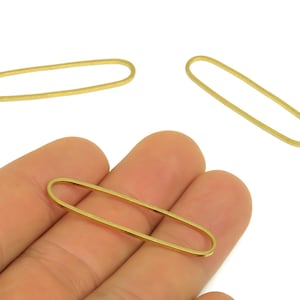 May include: Three gold-colored oval-shaped jewelry findings. One is held in a hand, while two others are scattered on a white background. The findings are made of a thin, metallic material.
