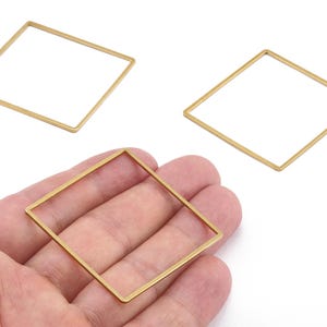 Puede incluir: Tres marcos cuadrados de metal dorado. Uno se sostiene en una mano, mientras que otros dos están colocados cerca. Los marcos tienen un diseño geométrico simple y probablemente se utilizan para la fabricación de joyas o manualidades.