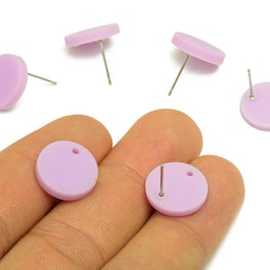 May include: A collection of light purple, round stud earrings with silver posts. The earrings are displayed on a hand against a white background. Each earring has a small hole near the post.