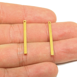 May include: Two gold-colored rectangular bar charms with a small hole at the top. The charms are held in a hand against a white background. These charms are suitable for jewelry making.
