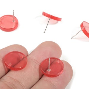 May include: Several translucent red disc-shaped stud earrings with silver posts are displayed. Two earrings are held in a hand, while others are scattered on a white surface. The earrings have a simple, minimalist design.