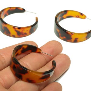Può includere: Tre orecchini a cerchio in tartaruga. Gli orecchini hanno un design ampio e curvo con un perno per la perforazione. Gli orecchini hanno un motivo di colori marrone scuro e ambra. Su sfondo bianco.
