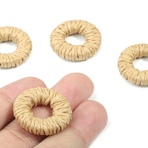 Könnte beinhalten: Nahaufnahme von mehreren beigefarbenen, kreisförmigen Raffiaperlen. Eine Perle wird in einer Hand gehalten, drei weitere liegen auf einer weißen Oberfläche. Die Donuts-förmigen Perlen bestehen aus eng gewickelten Naturfasern.