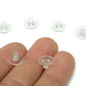 Pode incluir: Uma coleção de pequenas costas de brincos de plástico transparente. As costas dos brincos são redondas com uma base plana e um poste no centro. As costas dos brincos são mostradas na mão de uma pessoa.